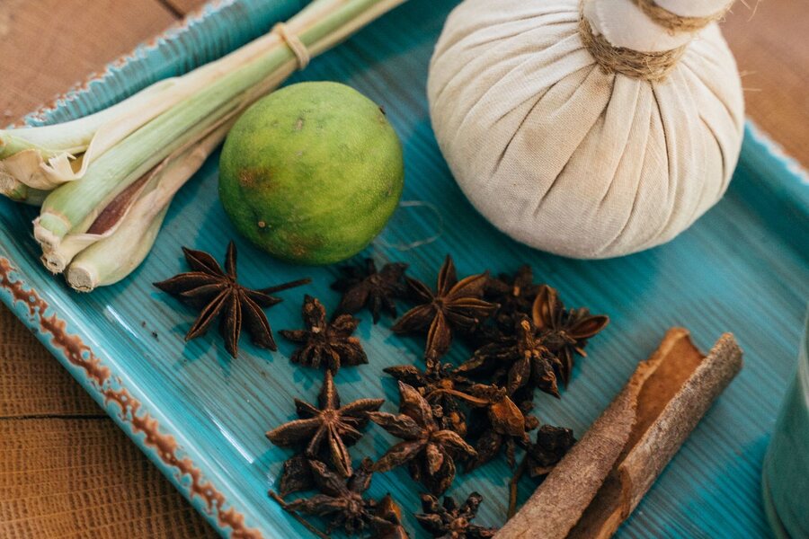 A Thai herbal compress ball beside lemongrass, a lime, star anise and cinnamon on a tray